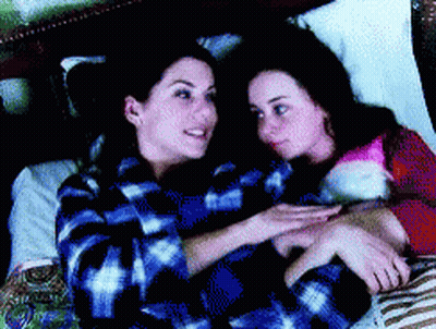 Two Women Laying On A Bed Looking At Each Other With One Wearing A Plaid Shirt GIF