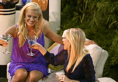 Two Women On Couch Drinking Cocktails And Smiling GIF