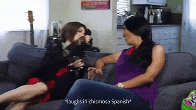 Two Women On Couch Saying Laughs In Chismosa Spanish GIF