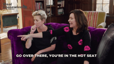 Two Women On Purple Couch Go Over There You're In The Hot Seat GIF