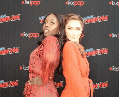 Two Women Pose For A Photo In Front Of A Comic Con Backdrop GIF
