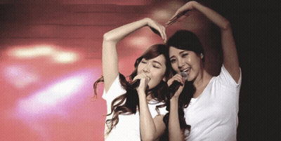 Two Women Singing Into Microphones And Making A Heart Shape GIF