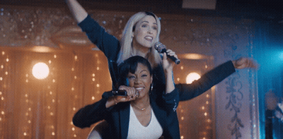 Two Women Singing Into Microphones With One Sitting On The Other 'S Shoulders GIF