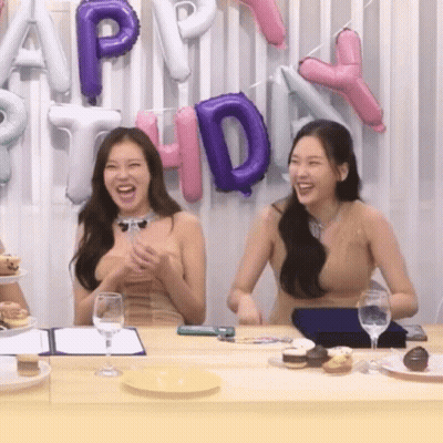 Two Women Sit At A Table In Front Of Balloons That Spell Out Happy Birthday GIF