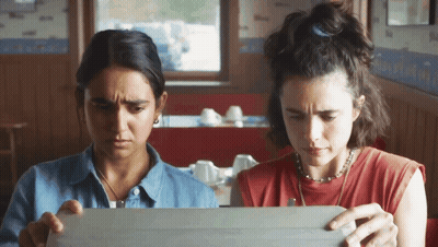 Two Women Sit At A Table Looking At A Box GIF