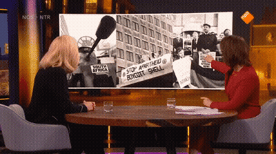 Two Women Sitting At A Table In Front Of A Screen That Says Nos On It GIF