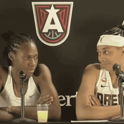 Two Women Sitting At Table With Microphones And Sign A GIF