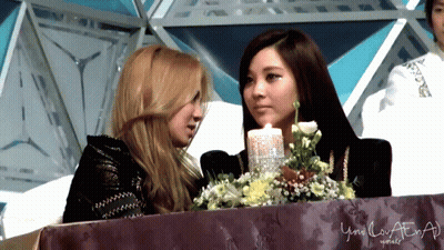 Two Women Sitting Next To Each Other In Front Of A Table With Flowers And A Sign That Says Your Love At GIF