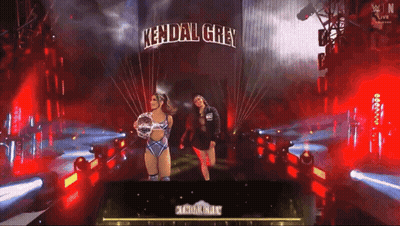 Two Women Standing In Front Of A Sign That Says Kendal Grey On It GIF
