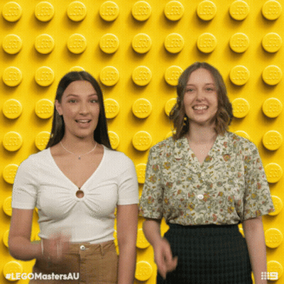 Two Women Standing In Front Of Yellow Lego Wall GIF