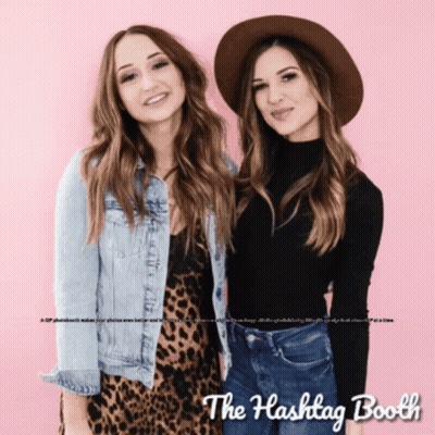 Two Women Standing Next To Each Other In Front Of A Pink Background With The Hashtag Booth Written On It GIF