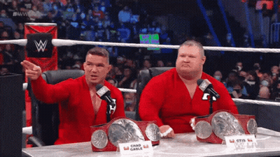 Two Wrestlers Sitting At A Table With A Sign That Says Chad Gable On It GIF