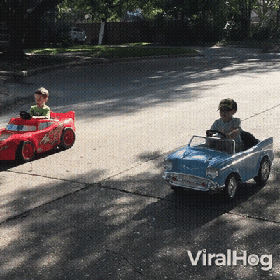 Two Young Boys Driving Toy Cars On Street GIF