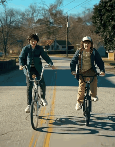 Two Young Boys Riding Bicycles Down A Street With One Wearing A T-Shirt That Says ' California ' On It GIF