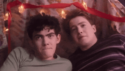 Two Young Men Are Laying Next To Each Other Under A Blanket With Christmas Lights . GIF
