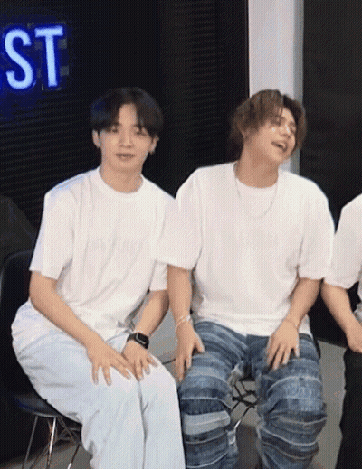 Two Young Men Are Sitting Next To Each Other In Front Of A Neon Sign That Says St GIF