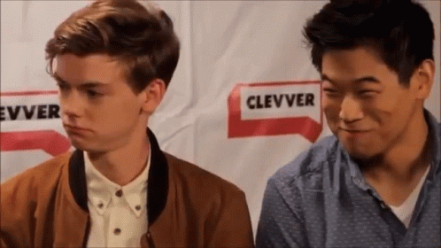 Two Young Men Are Sitting Next To Each Other In Front Of A Sign That Says ' Clever ' On It . GIF