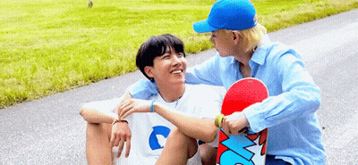 Two Young Men Are Sitting On The Side Of A Road Holding A Skateboard . GIF