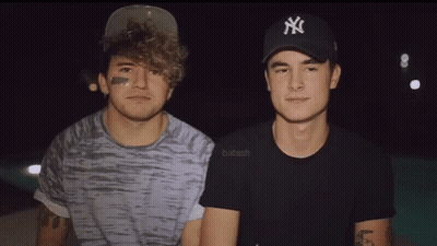 Two Young Men Are Standing Next To Each Other In A Dark Room . One Of The Men Is Wearing A Ny Hat . GIF