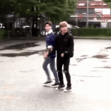 Two Young Men Are Walking Down A Street . GIF