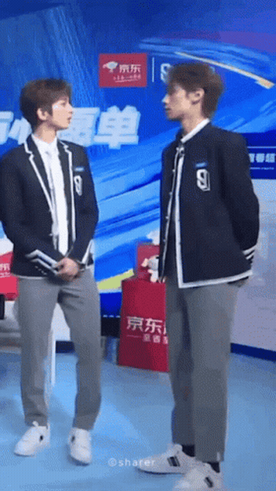 Two Young Men In Suits Are Standing Next To Each Other In Front Of A Large Screen . GIF