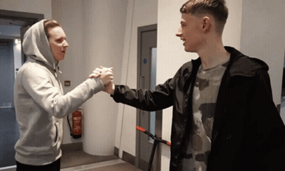 Two Young Men Shake Hands In A Hallway With A Fire Extinguisher On The Wall GIF