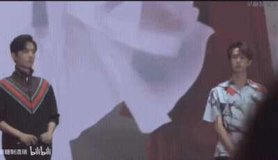 Two Young Men Standing Next To Each Other On Stage In Front Of Large Screen GIF