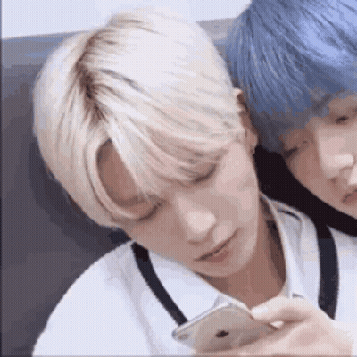 Two Young Men With Blonde Hair And Blue Hair Are Sitting Next To Each Other And Looking At A Cell Phone . GIF