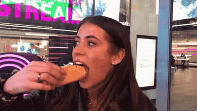 Woman Eating Sandwich In Front Of Stream Sign GIF