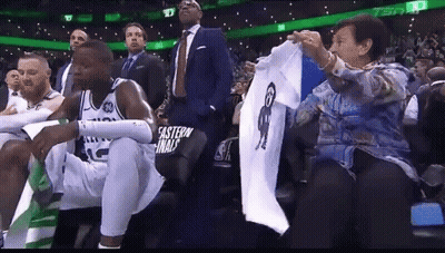 Woman Holding Shirt With Man Picture While Basketball Player Sits In Stands GIF