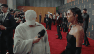 Woman In Black Dress Interviewed By Man In White Hat On Red Carpet GIF
