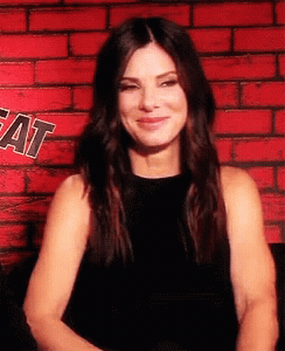 Woman In Black Dress Smiling In Front Of Red Brick Wall GIF