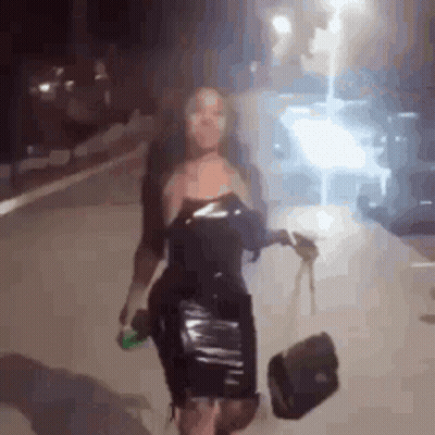 Woman In Black Dress Walking At Night Holding Purse And Beer GIF