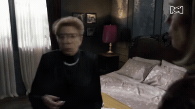 Woman In Black Shirt Standing In Front Of Bed With Pink Lamp GIF