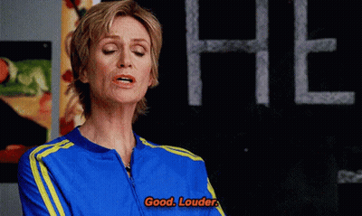 Woman In Blue And Yellow Jacket Says Good Louder In Front Of Chalkboard GIF