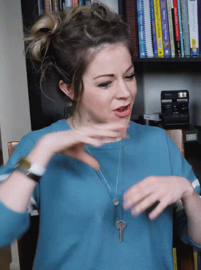 Woman In Blue Shirt With Key Necklace Sitting In Front Of Bookshelf GIF
