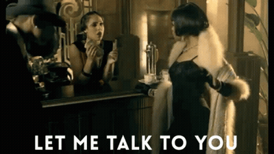 Woman In Fur Coat Talking To Man In Hat In Front Of Mirror GIF