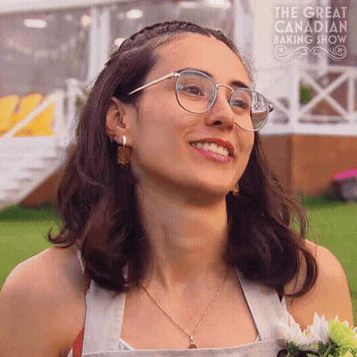 Woman in Glasses Smiling in Front of The Great Canadian Baking Show Sign