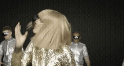 Woman In Gold Sequined Dress Dancing In Front Of Men In Silver Costumes GIF