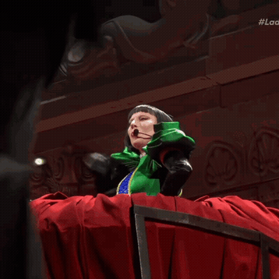 Woman In Green Dress And Black Gloves In Front Of Red Spiked Curtain GIF