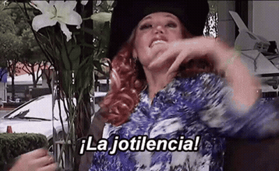 Woman In Hat And Blue Shirt Sitting Beside Flowers Says La Jotilencia GIF