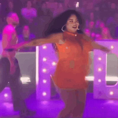 Woman In Orange Dress Dancing In Front Of Purple Letter GIF