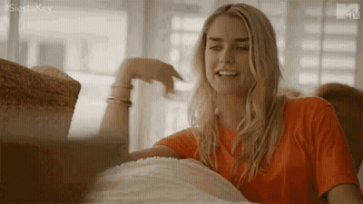 Woman In Orange Shirt Sitting On Couch Hand On Chin GIF