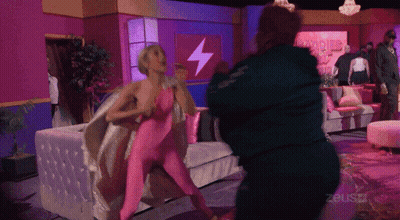 Woman In Pink Bodysuit Fighting Man In Green Jacket In Front Of Zeusz Sign GIF