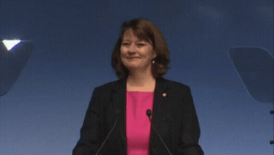 Woman In Pink Shirt And Black Jacket At Podium With Microphones GIF