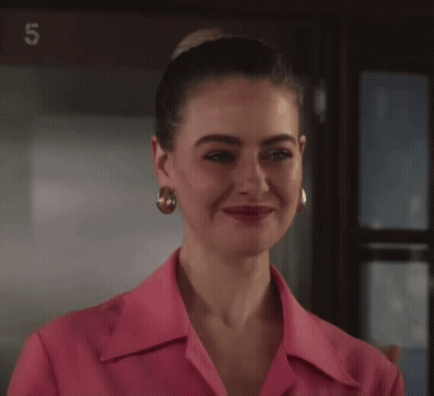 Woman In Pink Shirt And Gold Earrings Smiling In Front Of Sign Saying 5 GIF