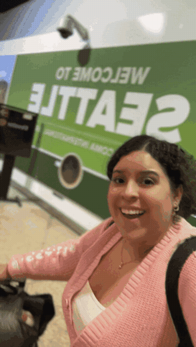 Woman In Pink Sweater Smiling In Front Of Green Sign Saying Elitta32 GIF