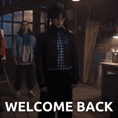 Woman In Plaid Shirt In Front Of Welcome Back Sign GIF