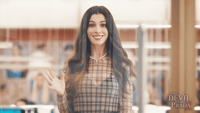Woman In Plaid Shirt Smiling And Waving In Front Of The Devil Wears Prada Sign GIF
