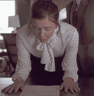 Woman In Polka Dot Blouse Kneeling At Desk Looking At Paper GIF